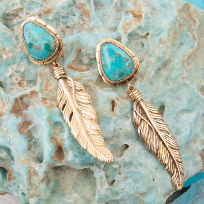 Gold feather earrings with turquoise stones on a textured stone background