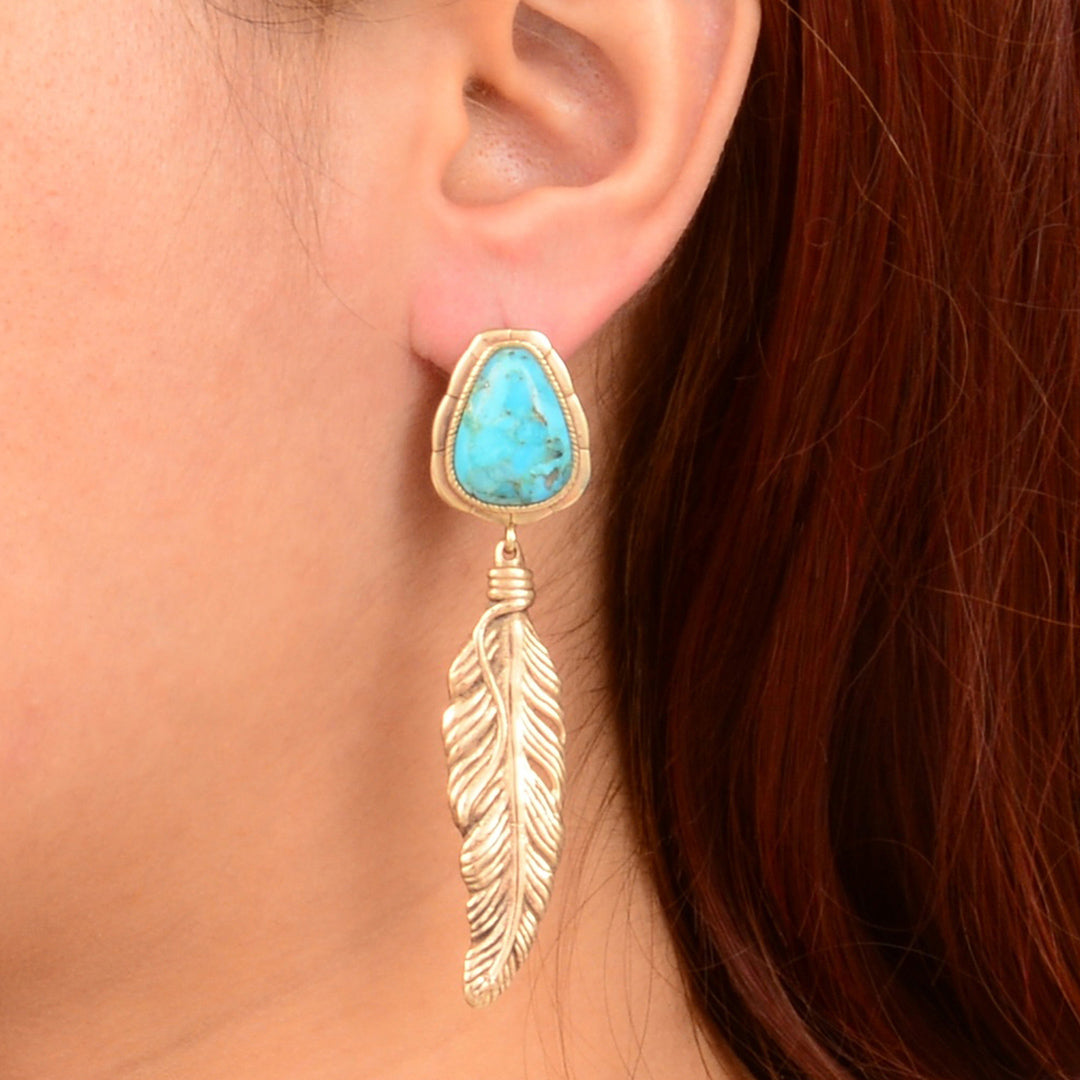 Gold feather earring with turquoise stone worn by a person