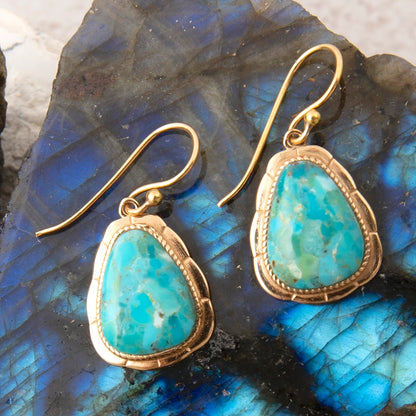 A pair of gold drop earrings with teardrop-shaped turquoise-blue stone inlays, displayed on a blue iridescent stone surface.