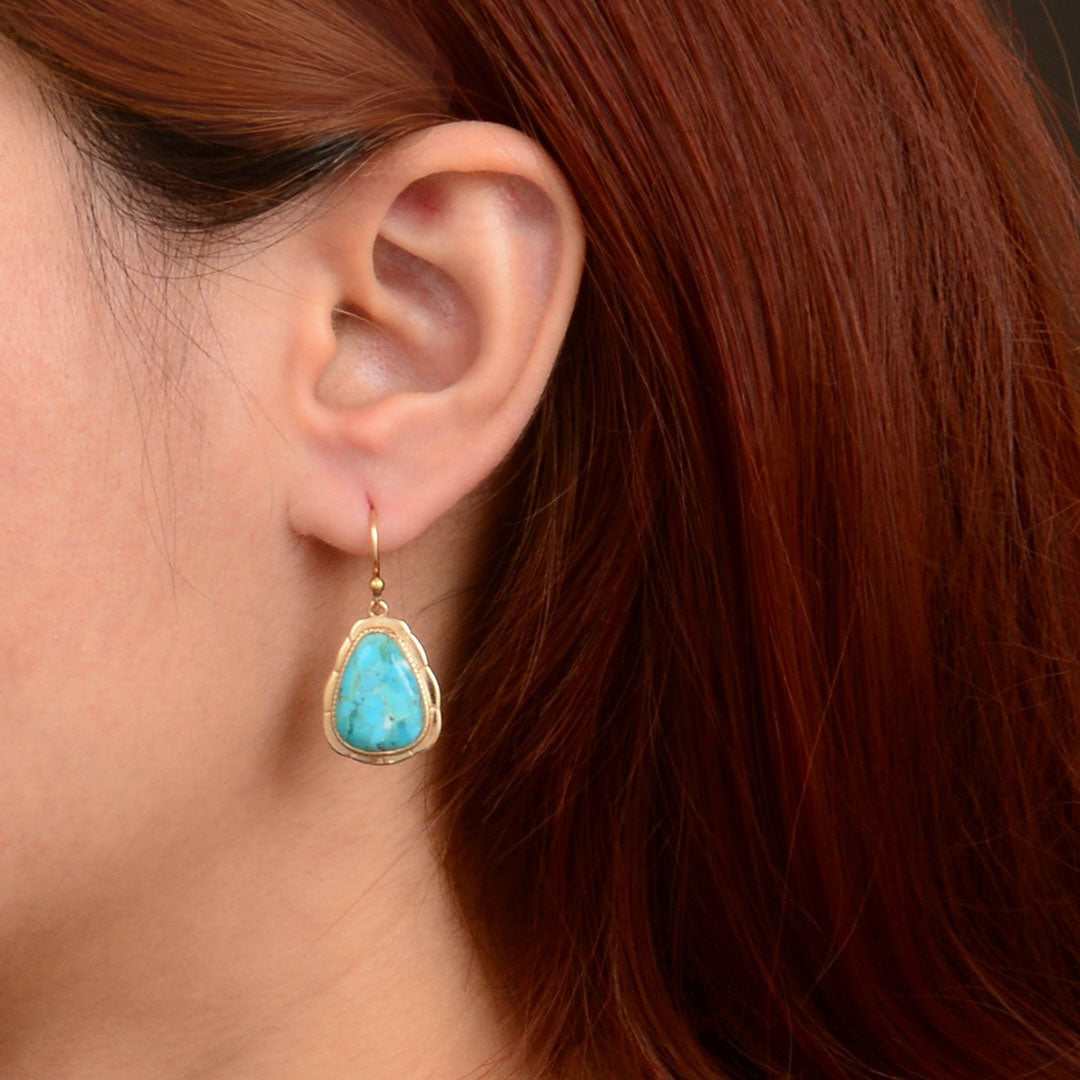 A close-up of a person's ear wearing a gold drop earring with a turquoise-colored teardrop-shaped stone.