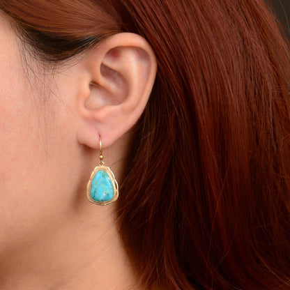 A close-up of a person's ear wearing a gold drop earring with a turquoise-colored teardrop-shaped stone.
