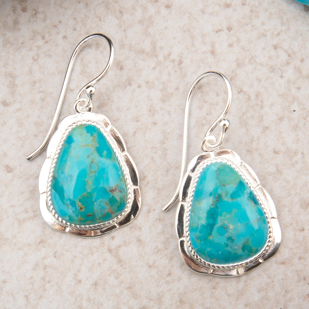 A pair of sterling silver dangle earrings featuring irregular turquoise-blue stone pendants with natural matrix veining, displayed on a light beige textured surface.