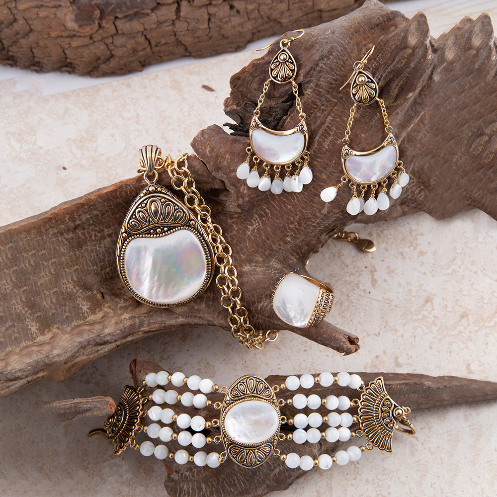A coordinated jewelry set displayed on a piece of driftwood over a light stone surface. The set includes a large pendant necklace, a ring, a bracelet, and matching earrings, all in antique gold-toned metal with iridescent white shell-like centers and small white bead drops.