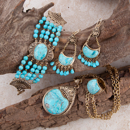 A jewelry set featuring turquoise-blue stone elements and ornate antique gold-toned metalwork, arranged on textured wood branches. The set includes a beaded bracelet, dangling earrings, a pendant necklace, and a ring.