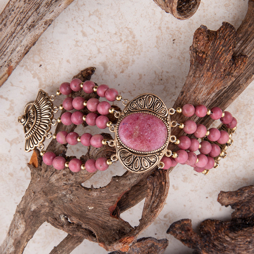 A multi-strand beaded bracelet featuring dusty pink beads, a central oval pink stone in an ornate antiqued gold-tone frame, and matching decorative end pieces, styled on driftwood branches over a light textured surface.