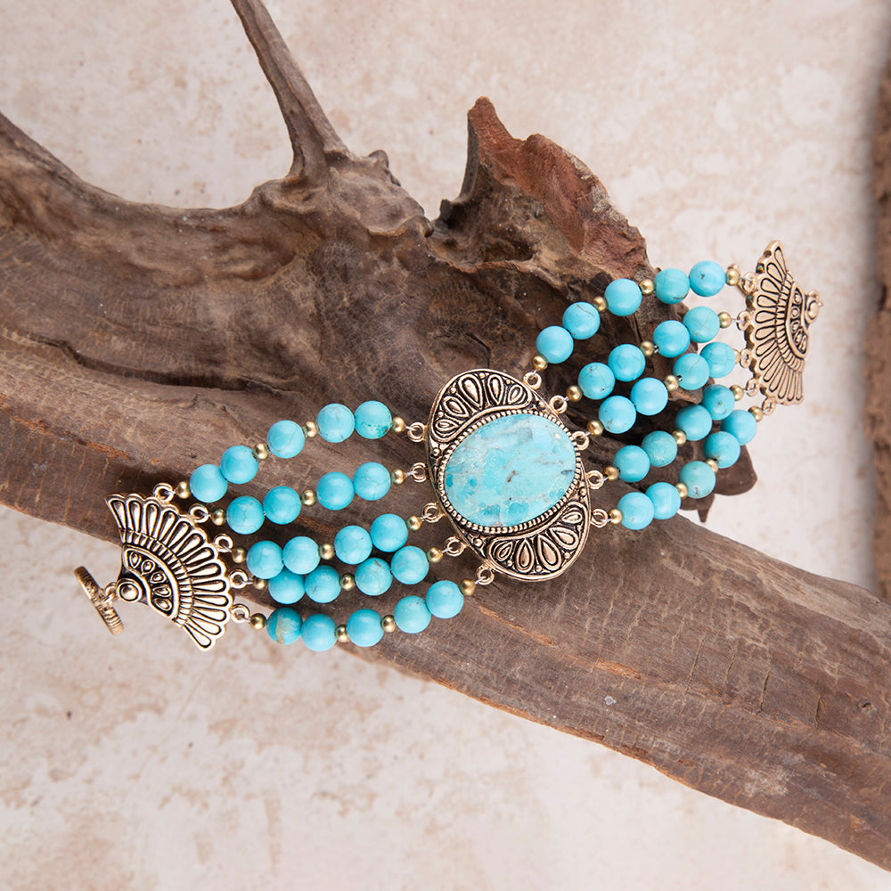 A multi-strand turquoise beaded bracelet with ornate antique gold-toned filigree details and a central oval turquoise stone, displayed on a piece of driftwood over a light beige textured surface.