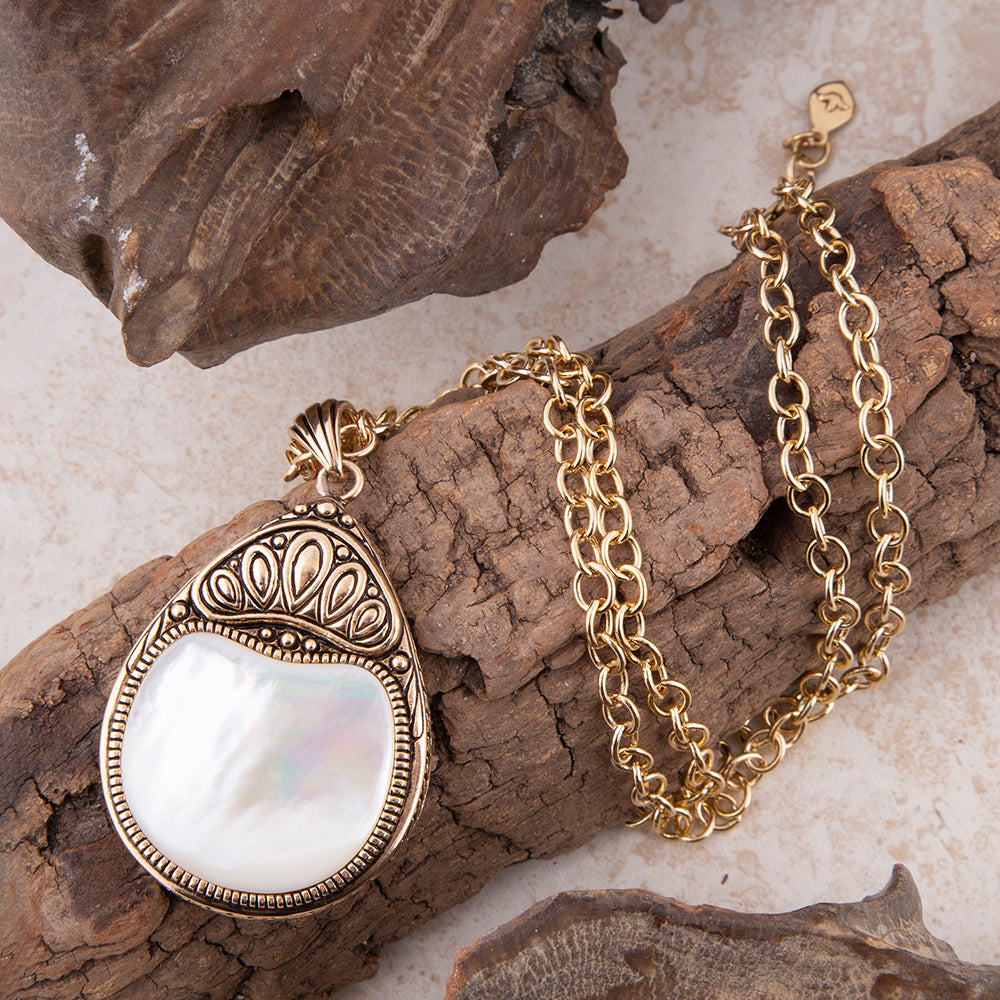 A gold pendant necklace with a teardrop-shaped white mother-of-pearl center and ornate filigree detailing, displayed on textured brown wood pieces over a light beige surface.