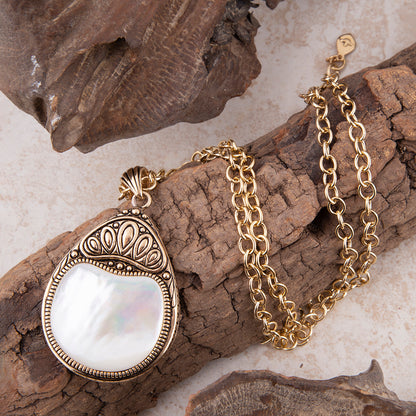 A gold pendant necklace with a teardrop-shaped white mother-of-pearl center and ornate filigree detailing, displayed on textured brown wood pieces over a light beige surface.