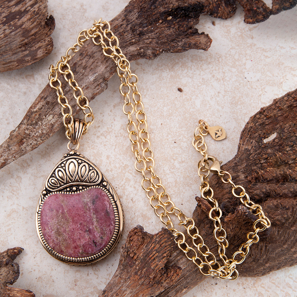 A gold chain necklace with an oval pendant featuring a pinkish-purple stone in a silver-toned bezel, styled on pieces of driftwood over a light textured surface.