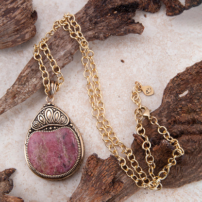 A gold chain necklace with an oval pendant featuring a pinkish-purple stone in a silver-toned bezel, styled on pieces of driftwood over a light textured surface.