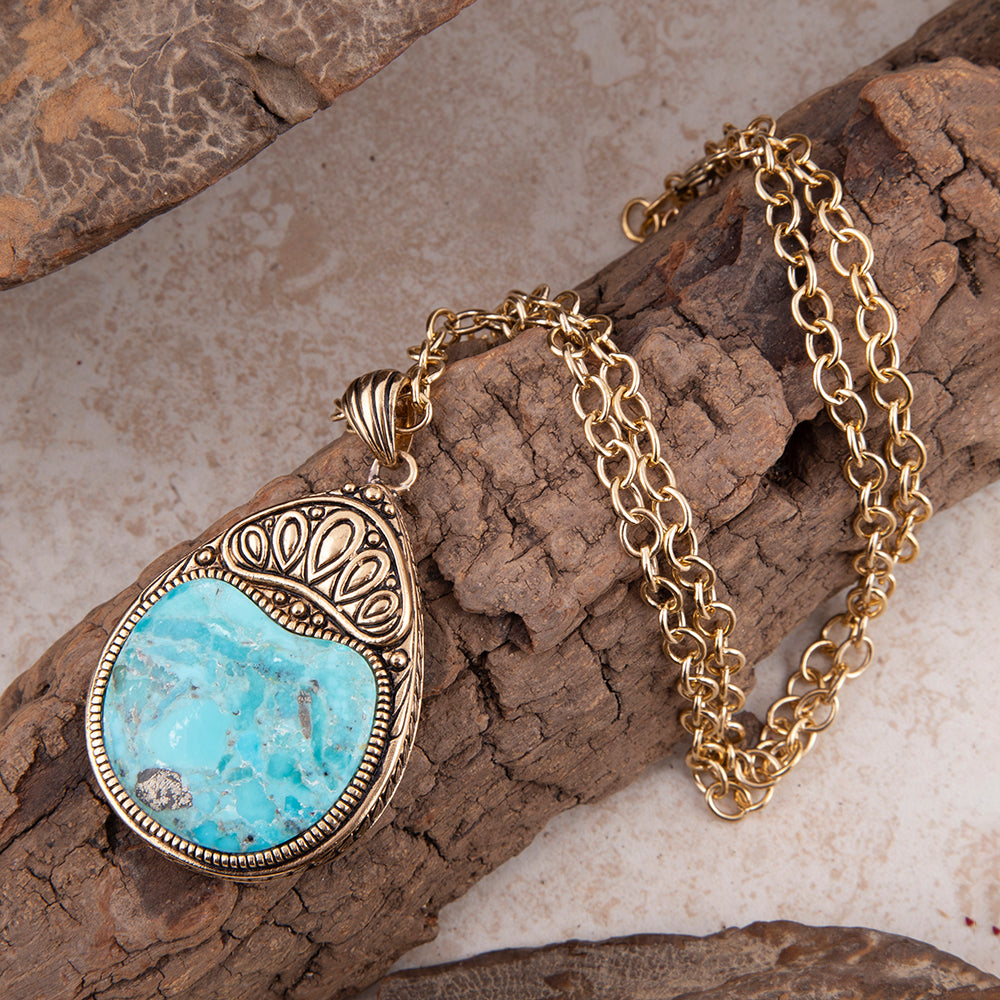 A turquoise and silver pendant necklace with a gold chain, styled on a piece of textured wood over a light stone surface.