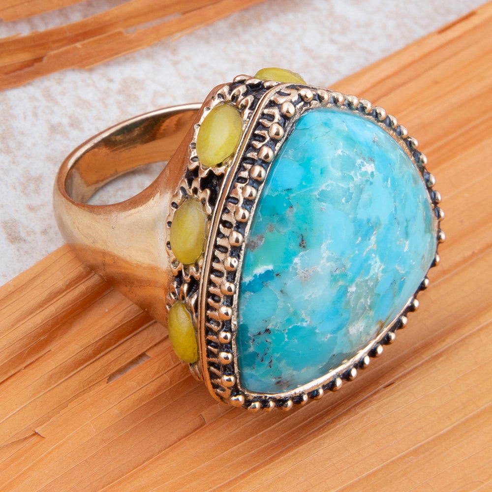 A silver-toned ring featuring a large, irregular-shaped light blue turquoise stone with natural marbling, accented by three small round yellow-green cabochon stones on the band, set against a bamboo background.