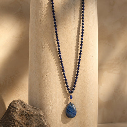 A long beaded necklace with dark blue beads and a teardrop-shaped blue stone pendant, displayed draped over a textured beige stone column.