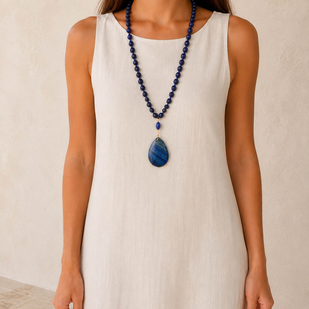 A woman wearing a long navy blue beaded necklace with a large teardrop-shaped blue stone pendant, paired with a sleeveless cream dress against a neutral beige wall.