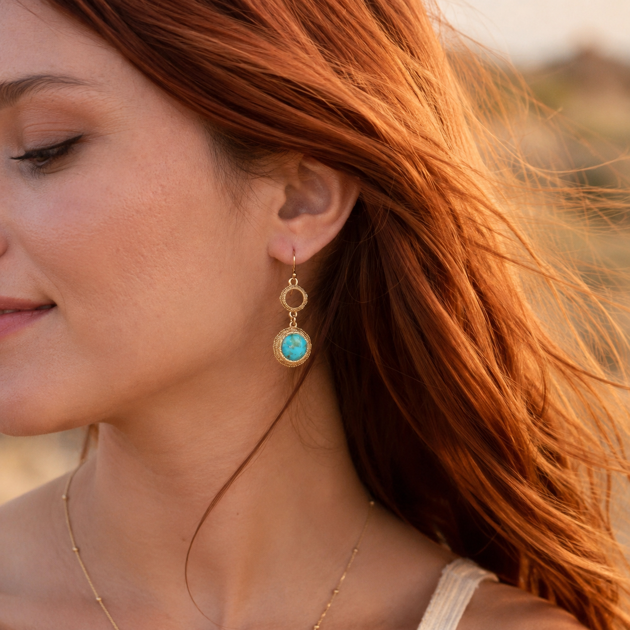 A close-up lifestyle shot of a woman with red hair wearing a gold drop earring with a round turquoise stone in a textured bezel setting.