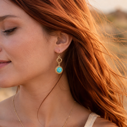 A close-up lifestyle shot of a woman with red hair wearing a gold drop earring with a round turquoise stone in a textured bezel setting.