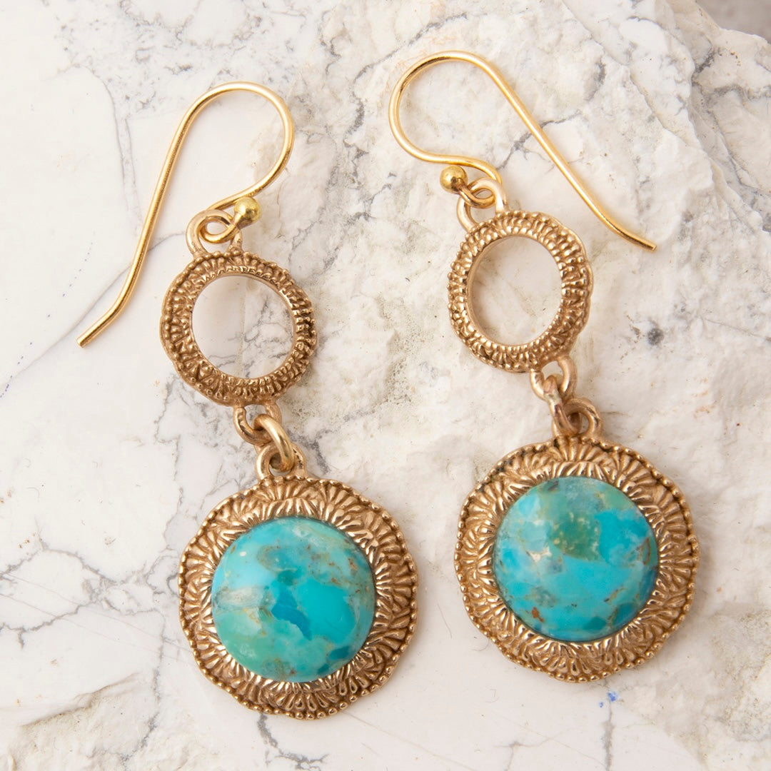 A pair of gold dangle earrings with textured circular hoops and oval turquoise stone drops, displayed on a white marble surface.