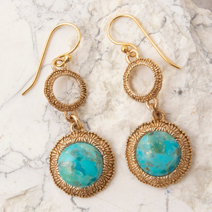 A pair of gold dangle earrings with textured circular hoops and oval turquoise stone drops, displayed on a white marble surface.
