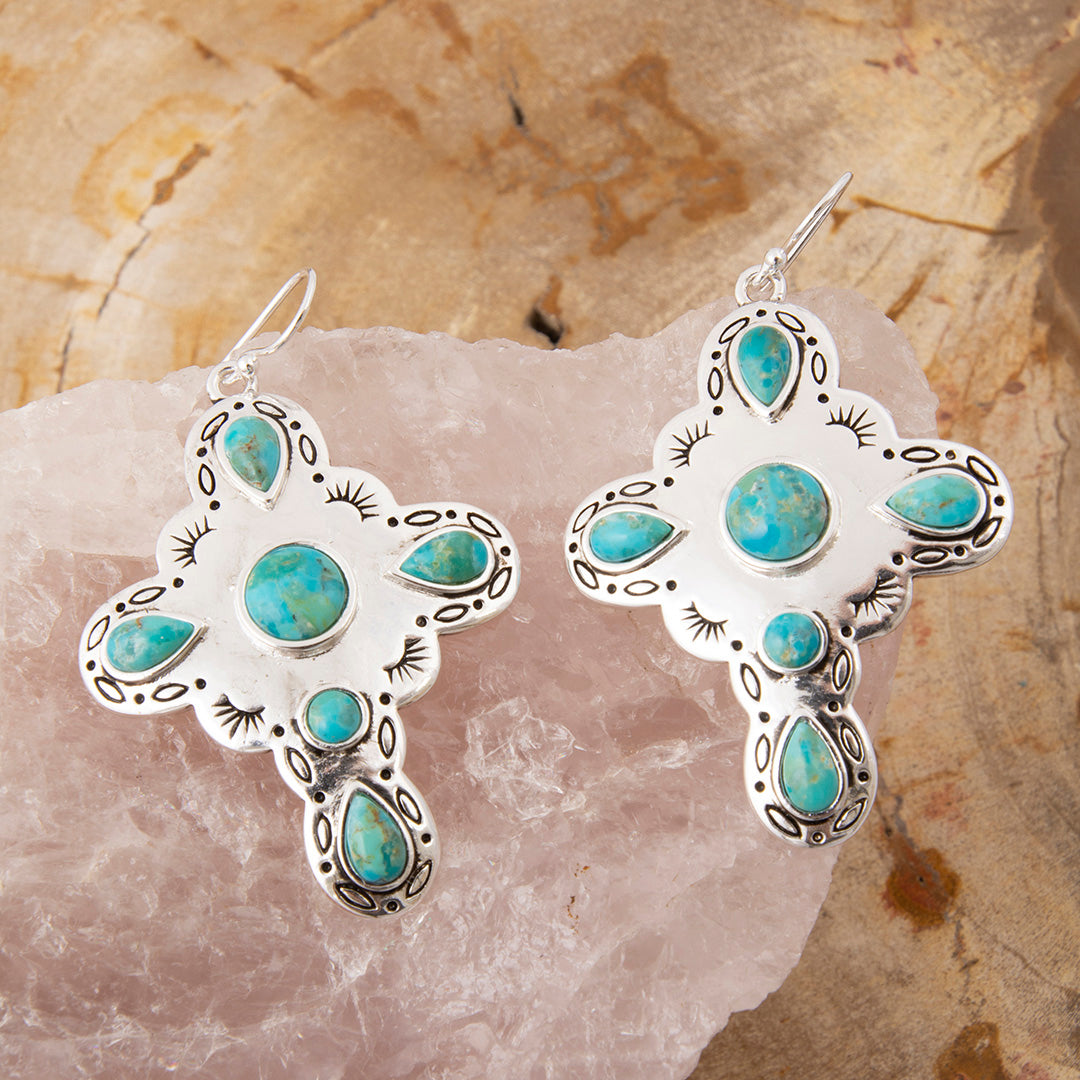 A pair of silver cross dangle earrings with turquoise stones, displayed on a pink crystal cluster over a textured tan stone background.