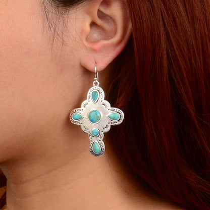 A close-up of a person's ear wearing a silver cross-shaped dangle earring with turquoise inlay.