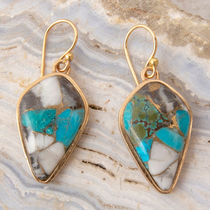 A pair of gold-tone drop earrings with irregular marquise-shaped turquoise, white, and gray stone inlays, displayed on a light gray agate slab background.