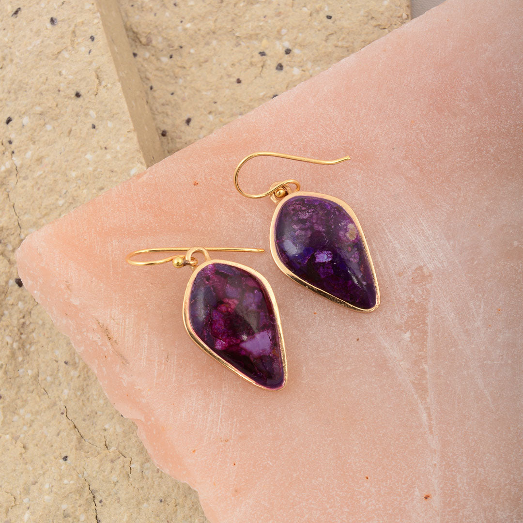 Purple teardrop earrings with gold hooks on a person's ear against a beige background