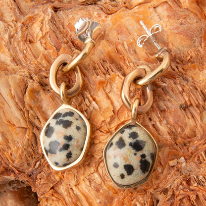 A pair of gold-tone dangle earrings with irregular beige and black spotted stone pendants, displayed on a textured brown wood background.