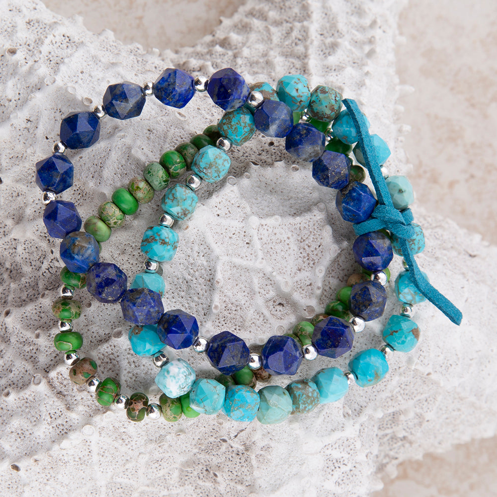 Three beaded wrap bracelets with blue, turquoise, and green stones, accented with silver beads and tied with a blue ribbon, displayed on a white textured lace background.