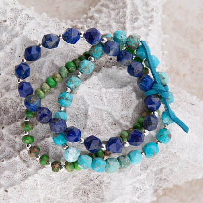 Three beaded wrap bracelets with blue, turquoise, and green stones, accented with silver beads and tied with a blue ribbon, displayed on a white textured lace background.