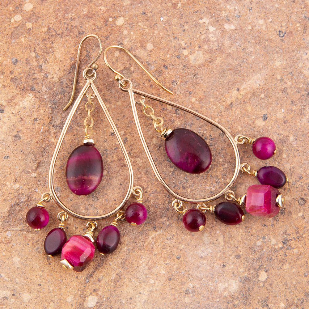 A pair of gold teardrop dangle earrings with large oval magenta beads and smaller round and faceted beads, displayed on a textured brown stone surface.