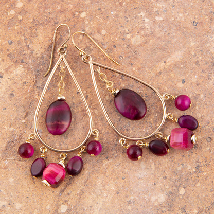 A pair of gold teardrop dangle earrings with large oval magenta beads and smaller round and faceted beads, displayed on a textured brown stone surface.