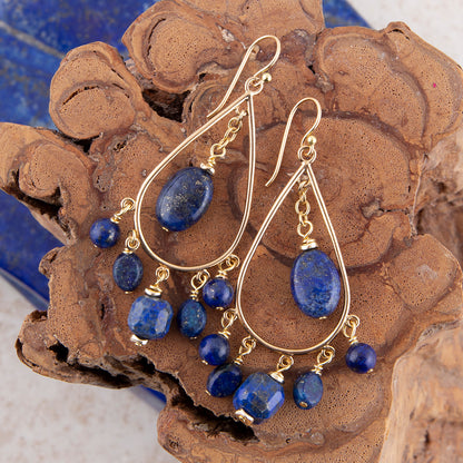 Gold teardrop dangle earrings with blue lapis lazuli stones, displayed on a textured brown wood background.