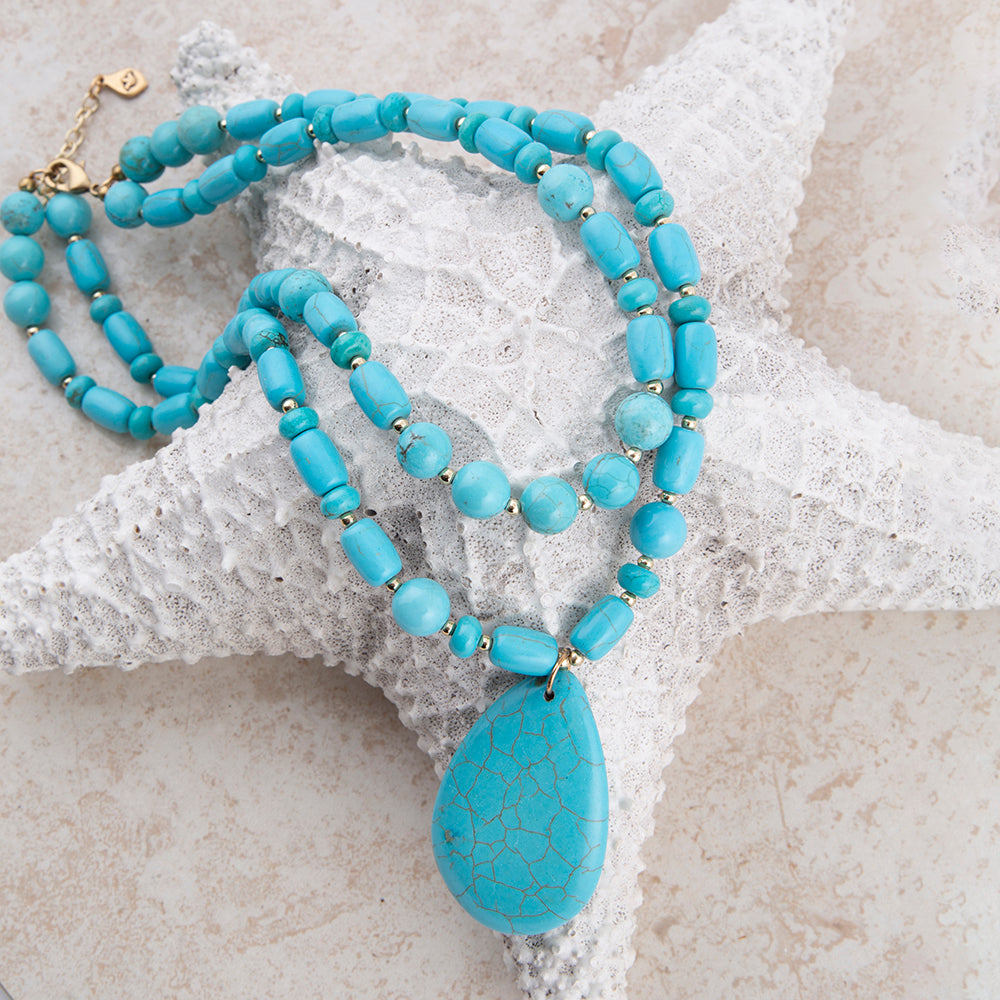 A turquoise-blue beaded necklace with a large teardrop-shaped crackled stone pendant, displayed on a white starfish prop against a light beige textured background.
