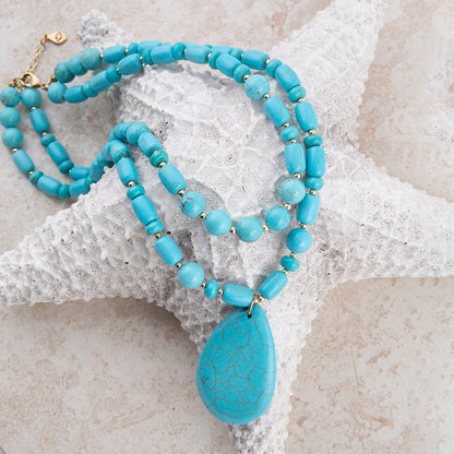 A turquoise-blue beaded necklace with a large teardrop-shaped crackled stone pendant, displayed on a white starfish prop against a light beige textured background.