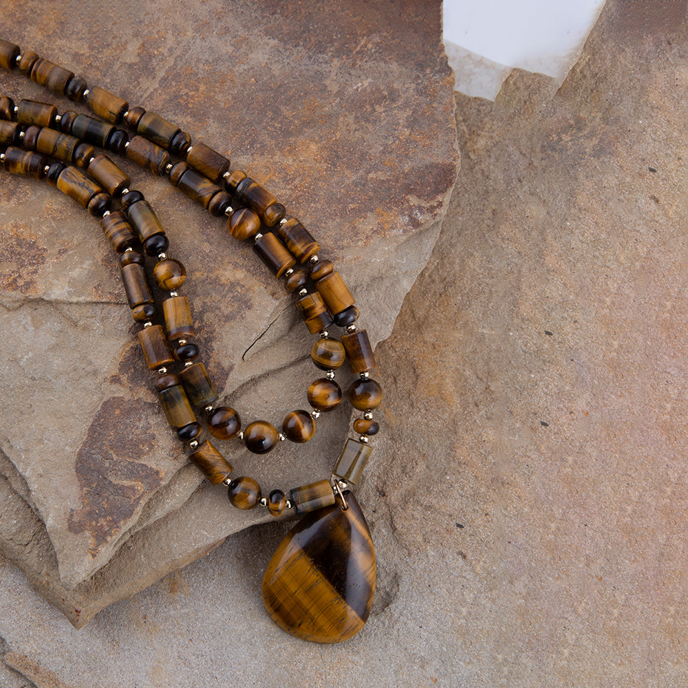 A beaded necklace made of polished tiger's eye beads in brown and gold tones, featuring a large teardrop-shaped pendant, displayed on textured stone slabs.