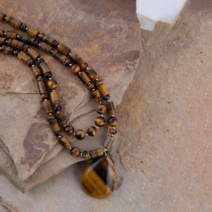 A beaded necklace made of polished tiger's eye beads in brown and gold tones, featuring a large teardrop-shaped pendant, displayed on textured stone slabs.