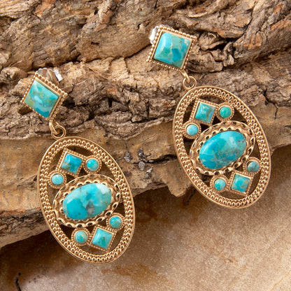A pair of gold-toned statement earrings featuring large oval and square turquoise cabochon stones, displayed on a textured piece of natural wood bark.