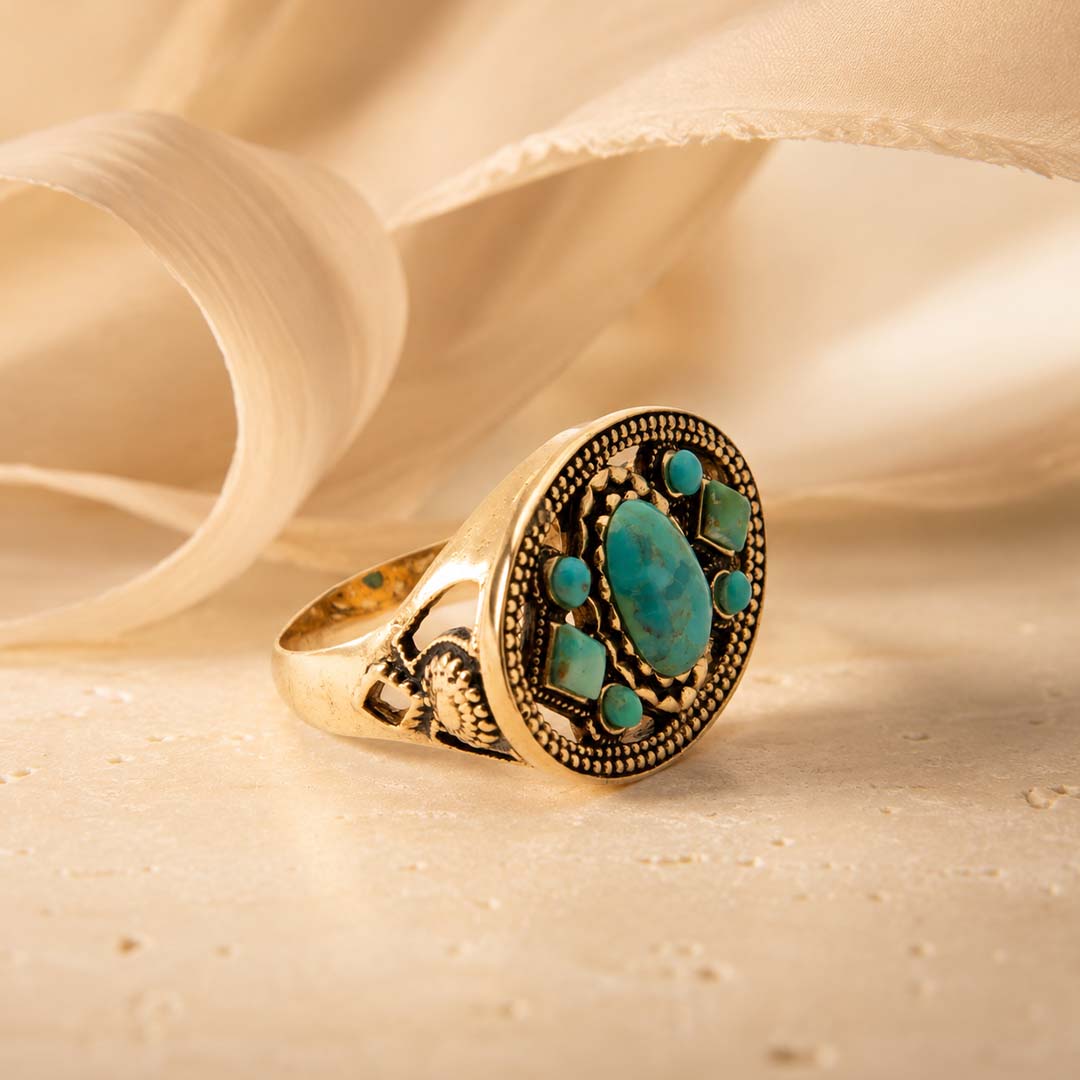 A gold signet ring with a large oval turquoise cabochon and smaller turquoise accents, styled on a textured beige surface with draped cream fabric in the background.