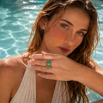 A woman with wet hair poses by a swimming pool, wearing a gold ring with a turquoise stone on her finger.