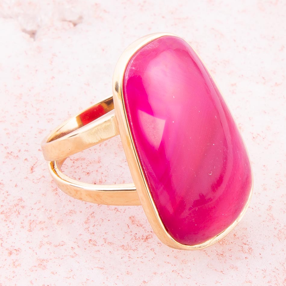 A gold ring featuring a large, smooth, oval-shaped magenta pink gemstone, photographed on a textured light pink surface.
