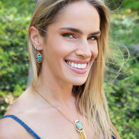 Woman wearing turquoise earrings and a necklace with a gold pendant, outdoors.