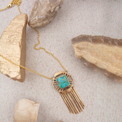 Gold necklace with a turquoise stone pendant on a textured surface with rocks.