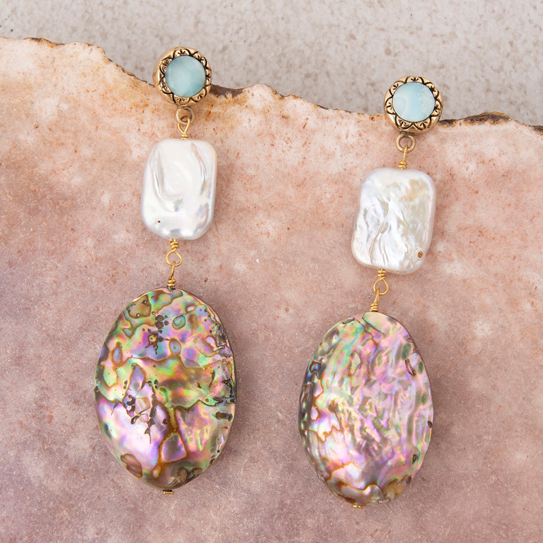 A pair of handmade dangle earrings featuring iridescent abalone shell drops, white baroque pearls, and gold-plated settings with light blue stones, displayed on a textured beige stone background.