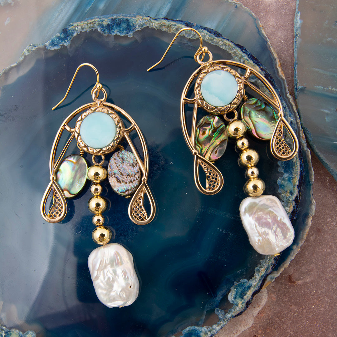 A pair of gold-tone dangle earrings with light blue oval stones, iridescent shell beads, and baroque pearl drops, displayed on a dark blue agate slice.