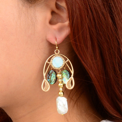 A close-up of a person's ear wearing a dangling gold earring with a light blue oval stone, two green iridescent shell pieces, and a white baroque pearl.