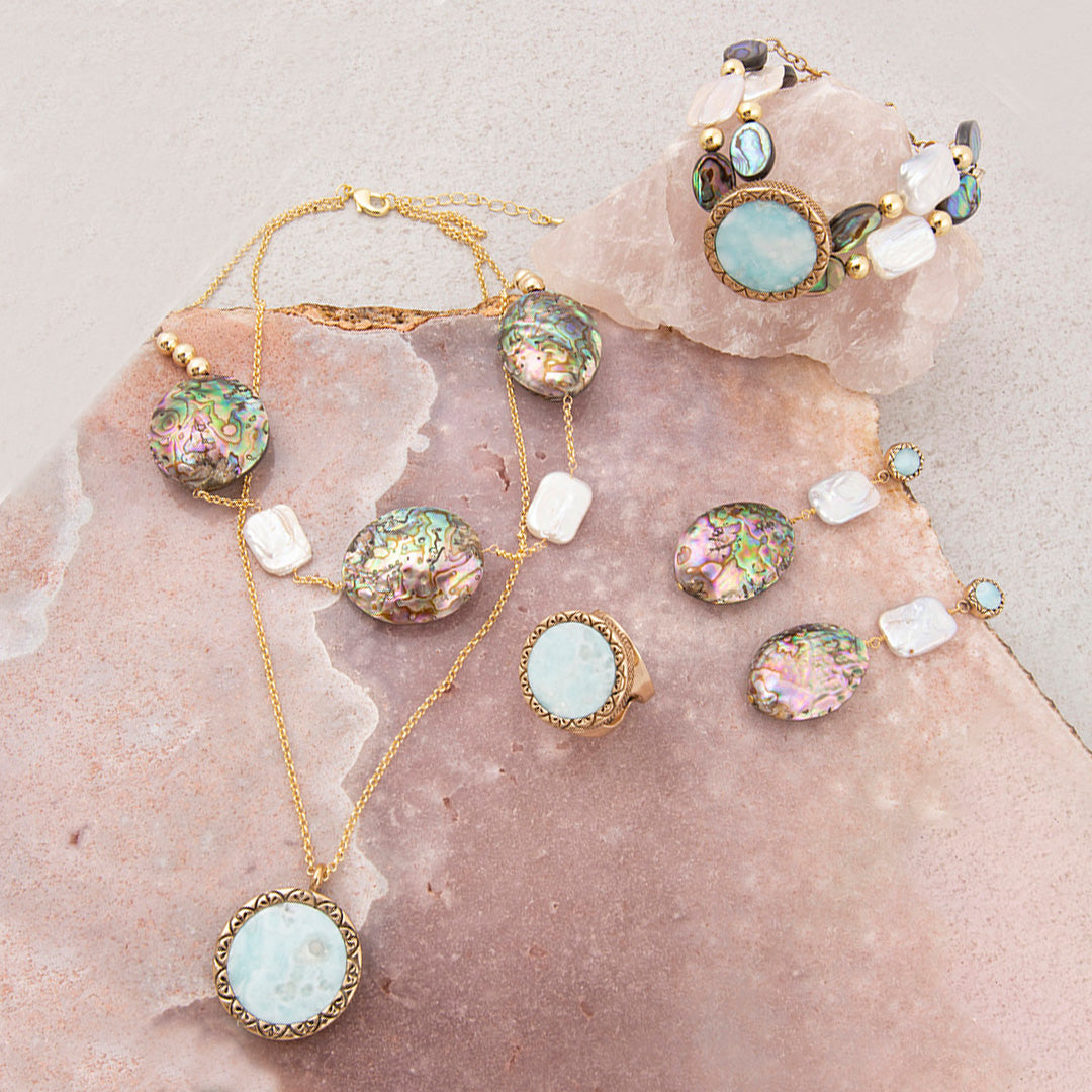 A coordinated jewelry set arranged on a pink stone slab, including a gold-tone necklace with a large iridescent oval pendant, matching earrings, a ring, and a bracelet. The jewelry features iridescent multicolor stones and pale translucent white stones in gold-toned settings.