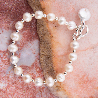 A beaded bracelet featuring large, lustrous white freshwater pearls separated by small silver spacer beads, finished with a silver toggle clasp, displayed on a textured pink and brown stone surface.