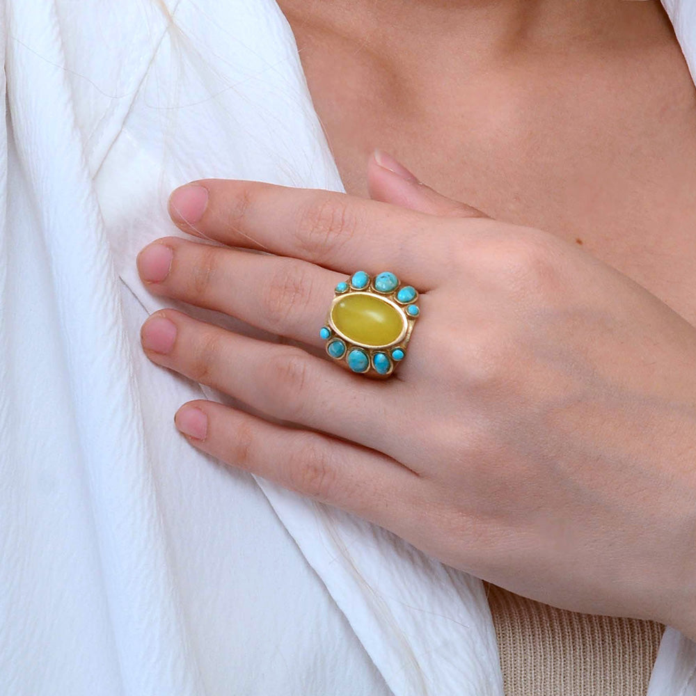 Hand wearing a gold ring with a yellow gemstone and turquoise stones on a white fabric background