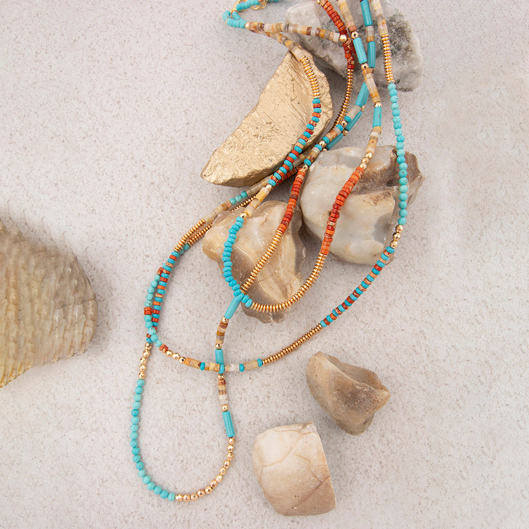 Beaded necklace with turquoise, gold, and orange beads on a sandy background with seashells.
