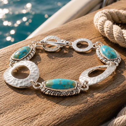 A silver-toned bracelet with two oval turquoise stones and textured circular links, displayed on a wooden surface near a rope and water.